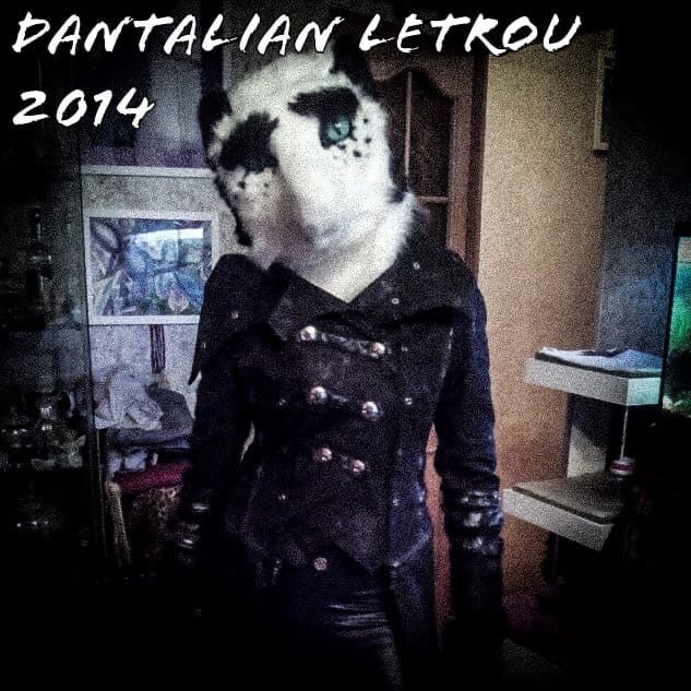 Фото с подписью «DANTALIAN LETROU 2014». Человек в белой фурри-голове (кот или собака с чёрными пятнами, зелёный глаз) в чёрном пальто с металлическими пуговицами и чёрных перчатках стоит в жилой комнате. Фото обработано в тёмном контрастном стиле.