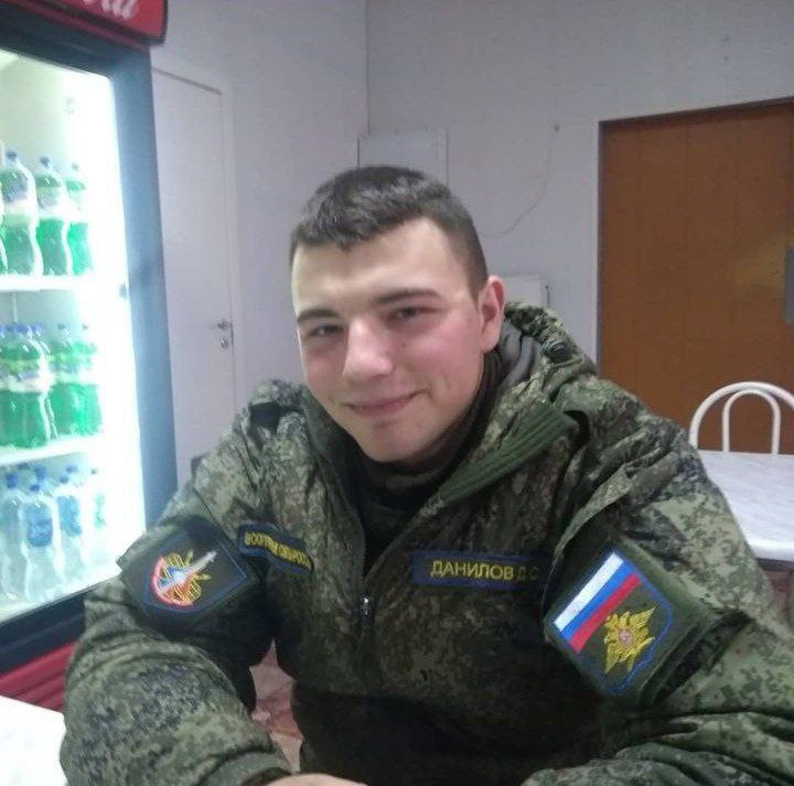Фото молодого мужчины в российской военной форме камуфляж, нашивка «ДАНИЛОВ Д.С.» и шеврон с флагом РФ. Сидит в столовой, на заднем плане холодильник с напитками.
