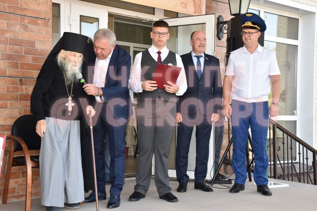Фото ВЧК-ОГПУ: групповое фото у входа в здание — схиархимандрит Илий с тростью, рядом организаторы в деловых костюмах, юноша с красной папкой и офицер в форме с синей фуражкой.