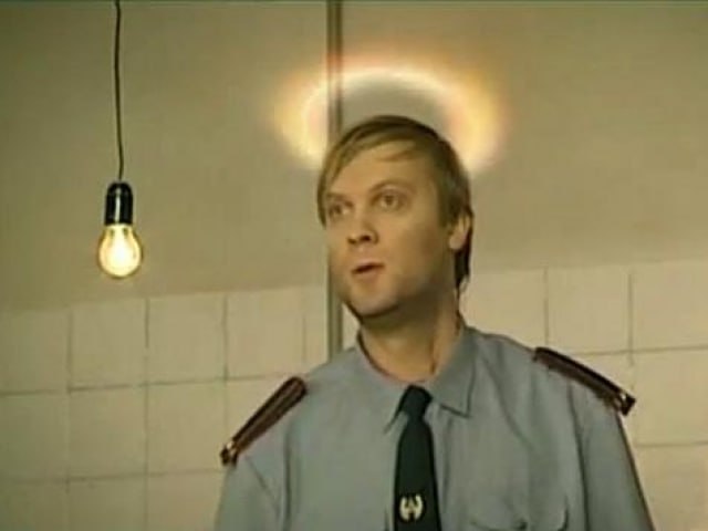 Кадр из советского/российского фильма или сериала: молодой светловолосый мужчина в форме милиционера стоит у белой кафельной стены, над его головой светится нимб (добавлен как мем). Сзади свисает лампочка.
