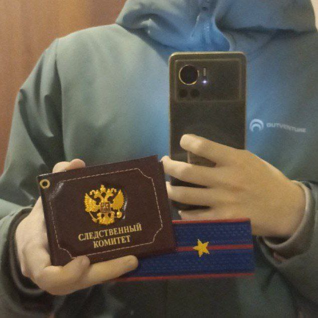 Мужчина в бирюзовой толстовке делает зеркальное селфи, держа в руках коричневое удостоверение «СЛЕДСТВЕННЫЙ КОМИТЕТ» с гербом РФ и что-то с погоном/звездой. Лицо не видно — закрыто телефоном.