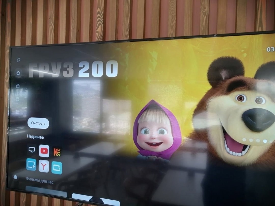 Фото Smart TV у деревянной стены. На экране — мультфильм «Маша и Медведь» (Маша в розовом платочке и медведь на жёлтом фоне). Поверх экрана наложен интерфейс: крупная надпись «ГРУЗ 200», кнопка «Смотреть», значки приложений (YouTube, Kinopoisk, Яндекс). Время 03:...