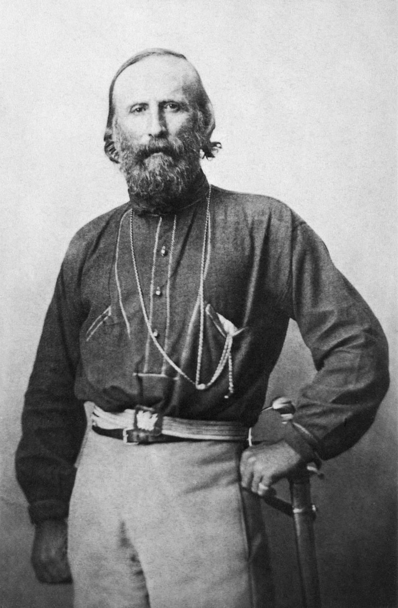 Чёрно-белая историческая портретная фотография. Мужчина средних лет с густой бородой и длинными волосами, в тёмной рубахе с несколькими цепочками через плечо. На поясе — сабля. Правая рука на рукояти, левая на поясе. Светлый студийный фон. Предположительно Джузеппе Гарибальди.