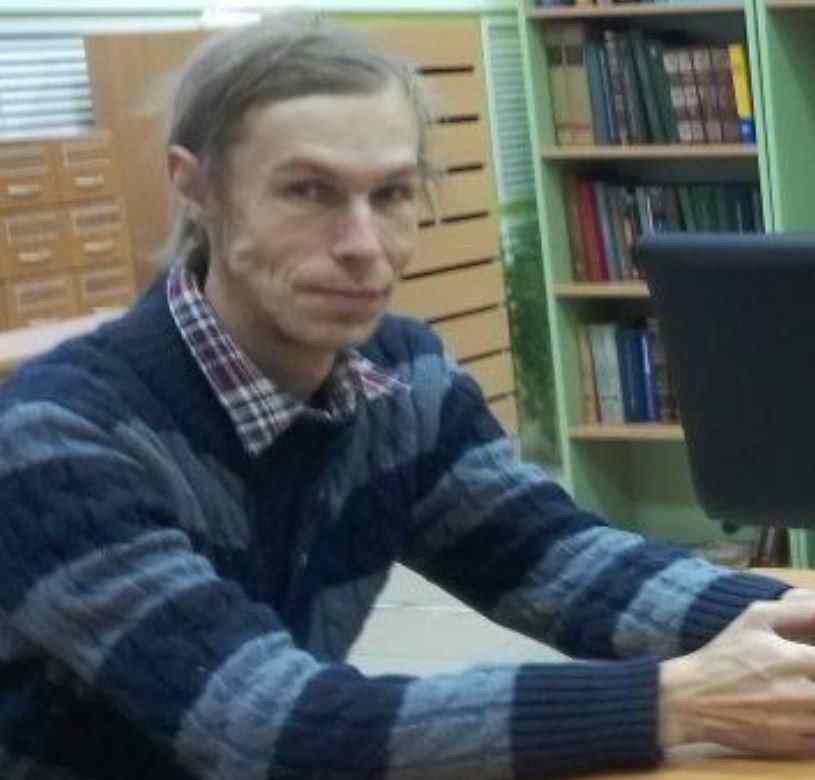 Фото мужчины, сидящего за столом у компьютера. Длинные светлые волосы зачёсаны назад, очень худое лицо с нестандартными чертами (широкий рот). Одет в синий полосатый свитер поверх клетчатой рубашки. На фоне — библиотечные стеллажи с книгами и картотечный шкаф.