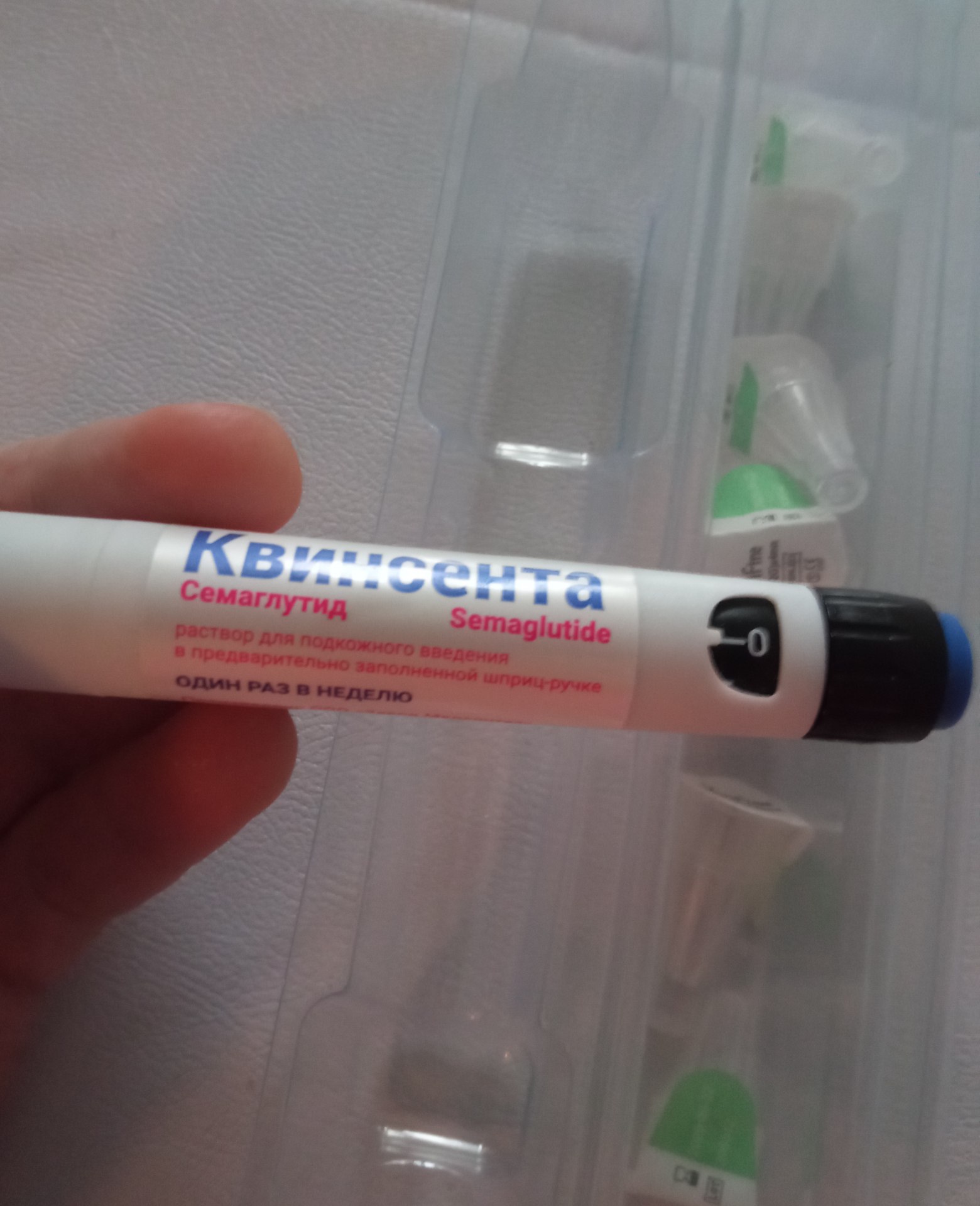 Фото: рука держит медицинскую шприц-ручку «КвинсентА — Семаглутид / Semaglutide» в белом корпусе с синим и красным текстом. Надпись: «Раствор для подкожного введения в предварительно заполненном шприц-ручке. ОДИН РАЗ В НЕДЕЛЮ». Доза на колесе установлена на 0. На фоне — упаковка с дополнительными иглами.