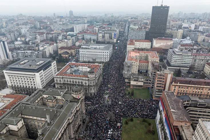 Аэрофотография центра Белграда в пасмурную погоду. Крупная площадь и прилегающие улицы полностью заполнены тысячами людей — массовая демонстрация против правительства Вучича (январь 2025). На заднем плане — городская застройка с высотными зданиями.