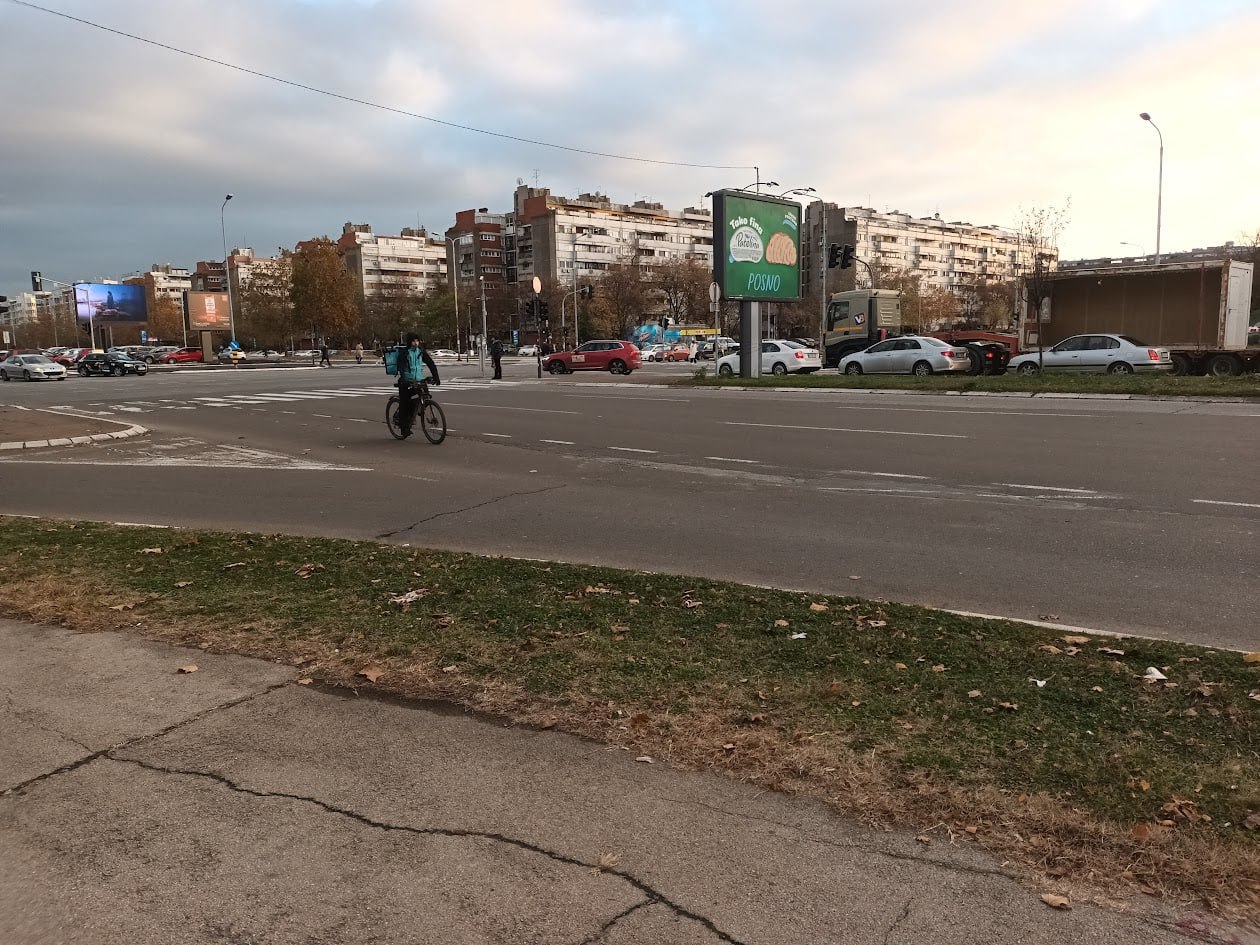 Городской перекрёсток: велосипедист едет по дороге, вдали жилые дома и рекламные щиты, осенние деревья без листьев, пасмурное небо.