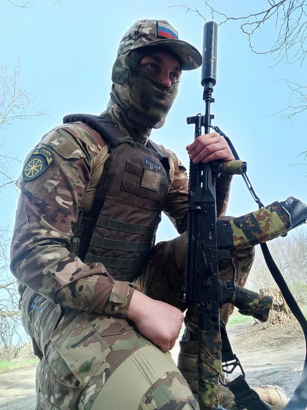 Военный в полном снаряжении: балаклава, мультикам-бронежилет, автомат; стоит облокотившись, на фоне деревьев.