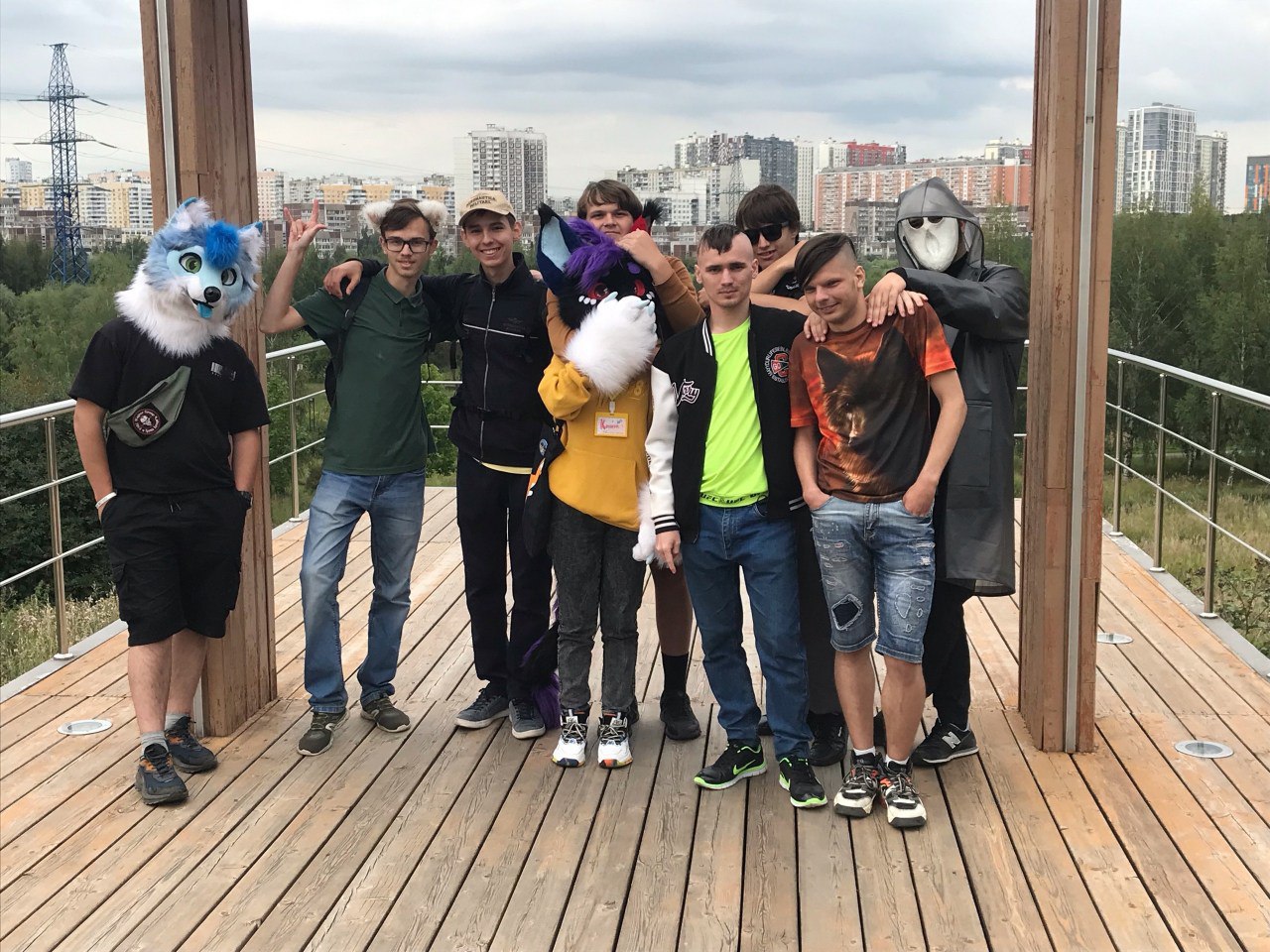 Группа примерно из восьми молодых людей фотографируется на деревянной открытой смотровой площадке; один из них в фурсьюте голубого волка, на заднем плане городские кварталы.