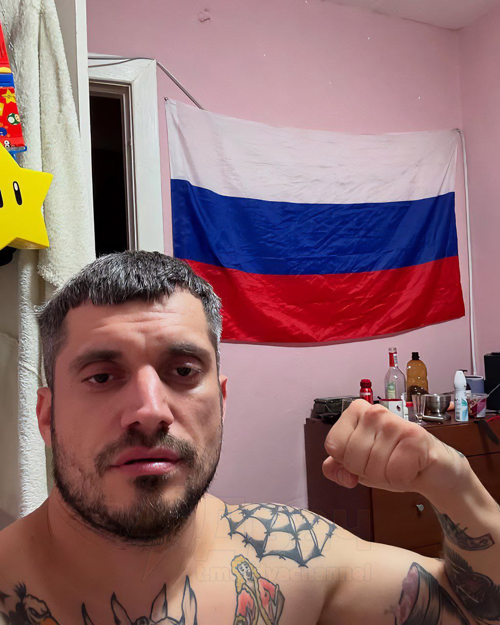 Мужчина без рубашки с татуировками на груди (паутина) и плечах, делает «козу» кулаком; на розовой стене за ним висит флаг России.