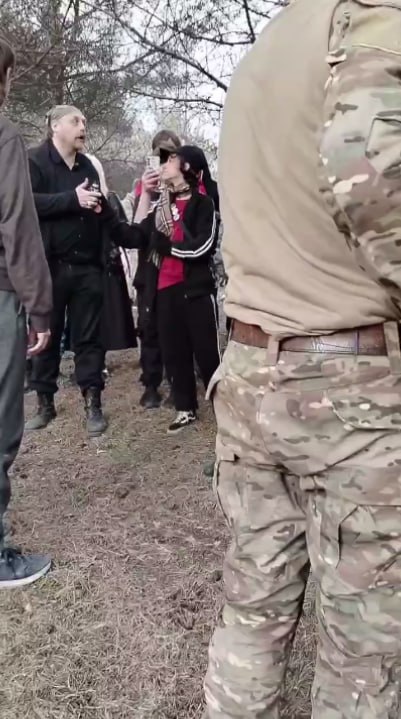 Фото в парке: несколько человек стоят на грунтовой дорожке, в центре — два человека в чёрной одежде разговаривают, справа — человек в камуфляже стоит спиной к камере.