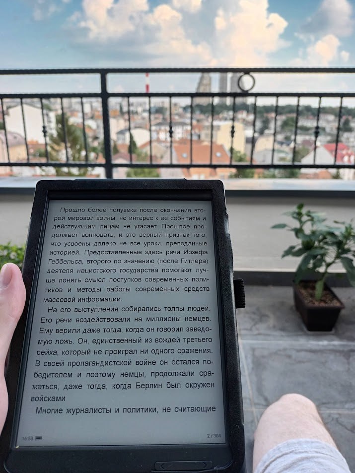 Рука держит электронную книгу (e-ink ридер) на фоне летнего городского вида с балкона; на экране текст о речах Геббельса, деятеля нацистского государства, и методах работы современных средств массовой информации.