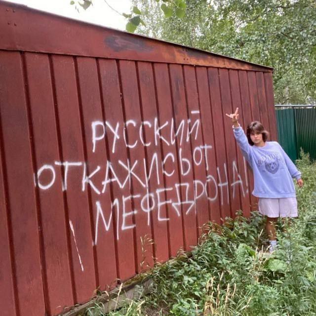 Девушка стоит у красного деревянного забора с граффити «Русский, откажись от мефедрона!»