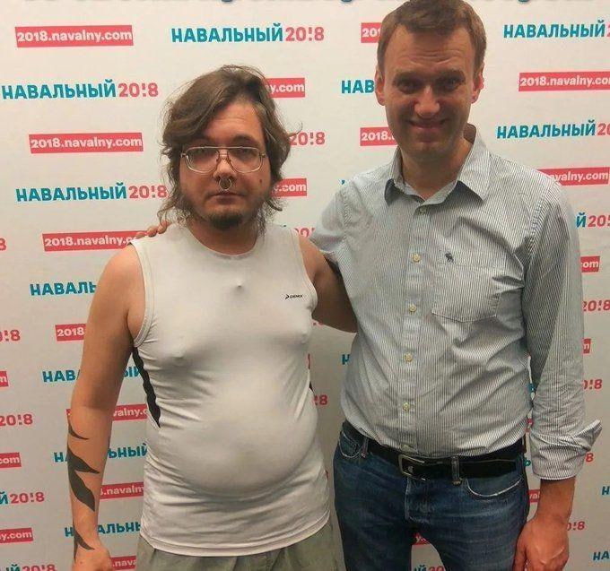 Фото (4/4): тот же мужчина без тату, в белой майке, стоит рядом с Алексеем Навальным на фоне баннеров «Навальный 2018».