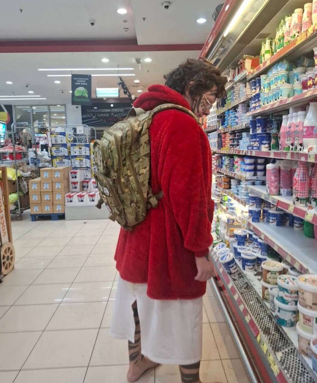 Фото из супермаркета в Подгорице: мужчина спиной к камере в красном плюшевом пальто и камуфляжном тактическом рюкзаке стоит у полок с молочными продуктами. Идентифицирован как Фосса (Shakti Lucidia, Артём Ветров).
