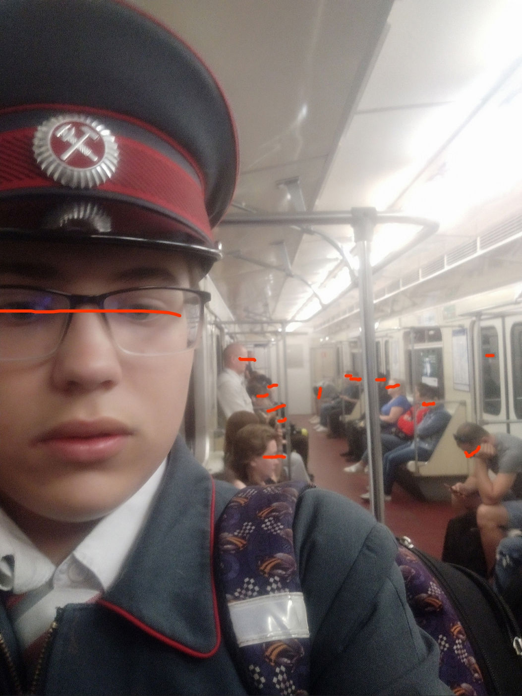 📷 Молодой человек (~17–18 лет) в форме железнодорожника (тёмно-серый мундир, фуражка с кокардой, очки, нашивка-бейдж на груди); в вагоне метро; лицо скрыто горизонтальной оранжевой полосой; на заднем плане пассажиры, поручни, двери вагона.