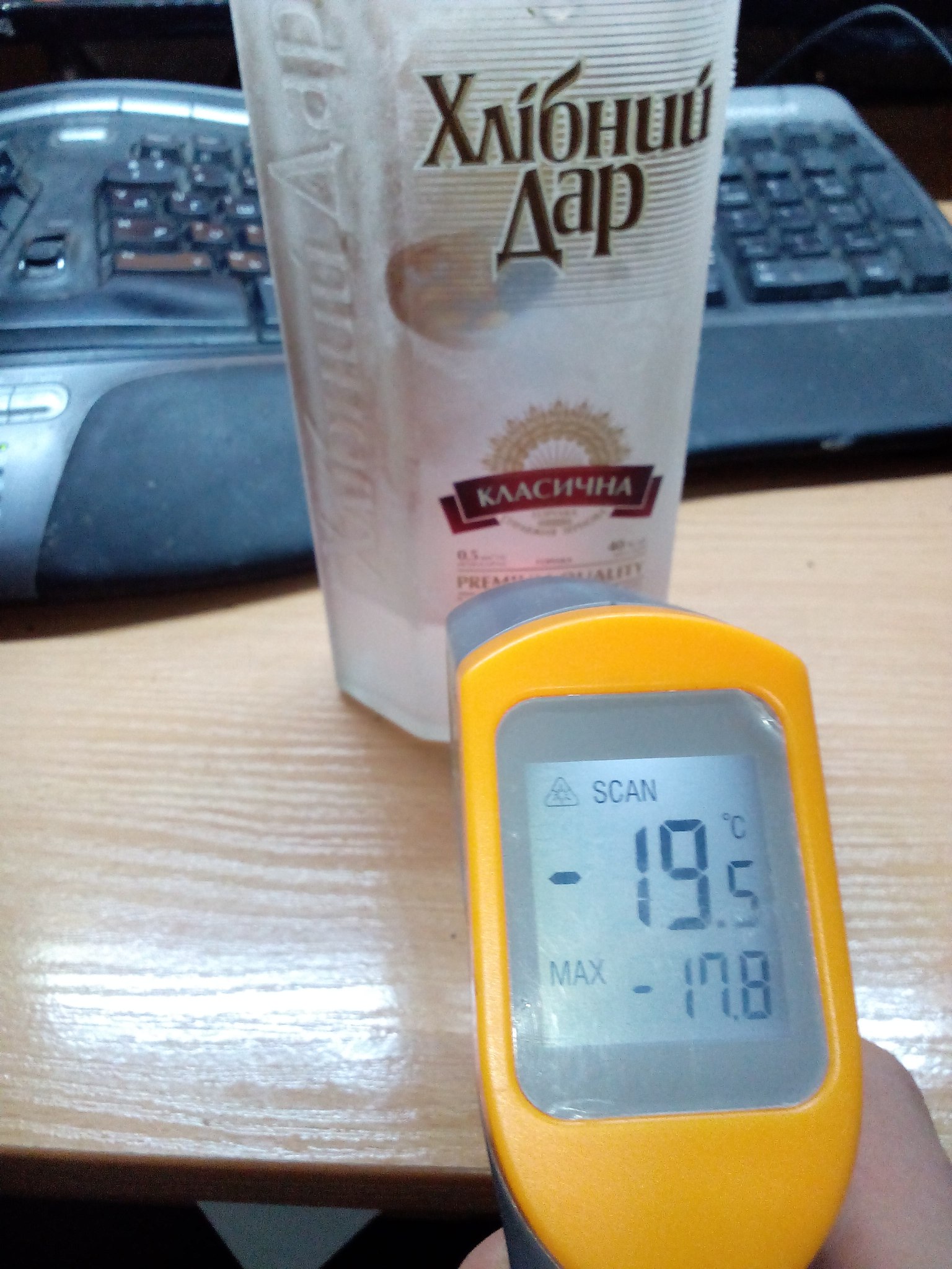 Жёлтый пирометр в руке показывает −19.5°C (MAX −17.8°C); на столе бутылка украинской водки «Хлібний Дар Класична 0.5 л»; на заднем плане два ноутбука