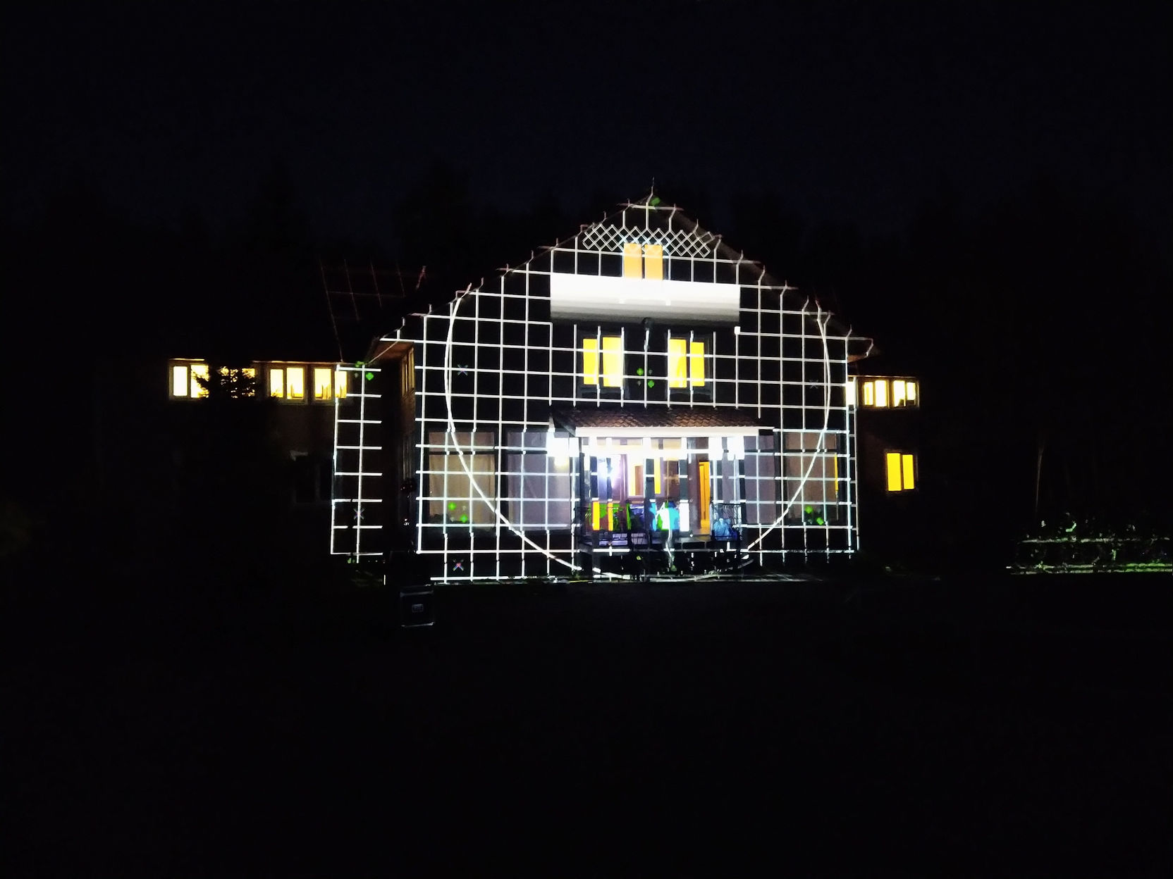 Ночная фотография видеомэппинга на фасаде двухэтажного здания с двускатной крышей: на стену спроецирована сетка из белых квадратов с наложенной белой окружностью; окна светятся жёлтым; под навесом крыльца разноцветные подсветки; художественная световая инсталляция