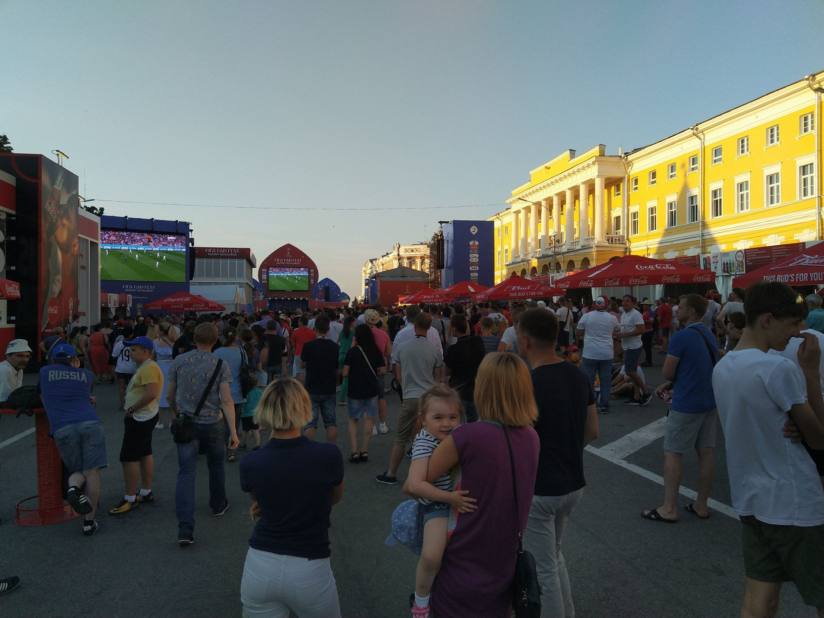 Широкая городская площадь в сумерках, заполненная людьми: слева крупный экран фан-зоны Чемпионата мира по футболу с трансляцией матча; справа — крупное жёлтое классическое здание; на переднем плане женщина с ребёнком на руках смотрит на экран
