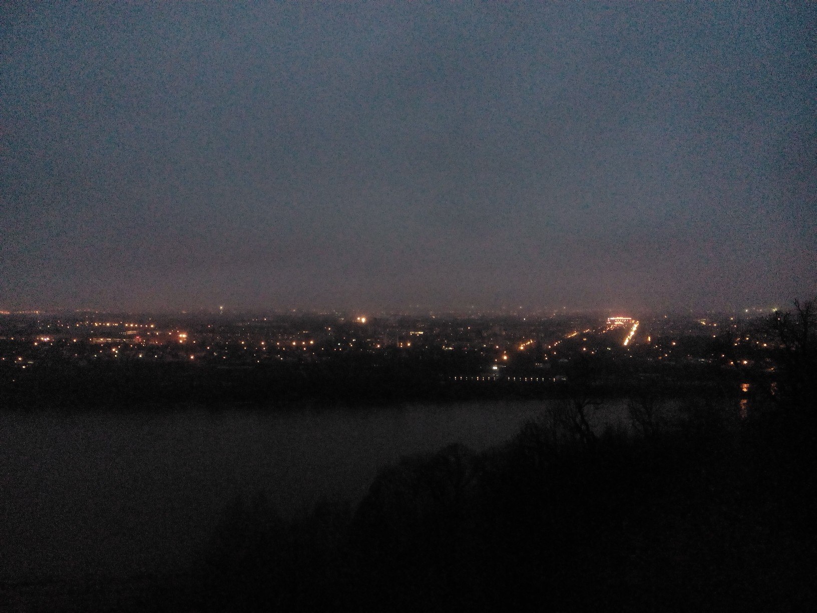 Ночная панорама с высокого берега: тёмная река внизу, на другом берегу — цепочки жёлтых огней вдоль дороги и жилых кварталов, тёмное небо с низкими облаками