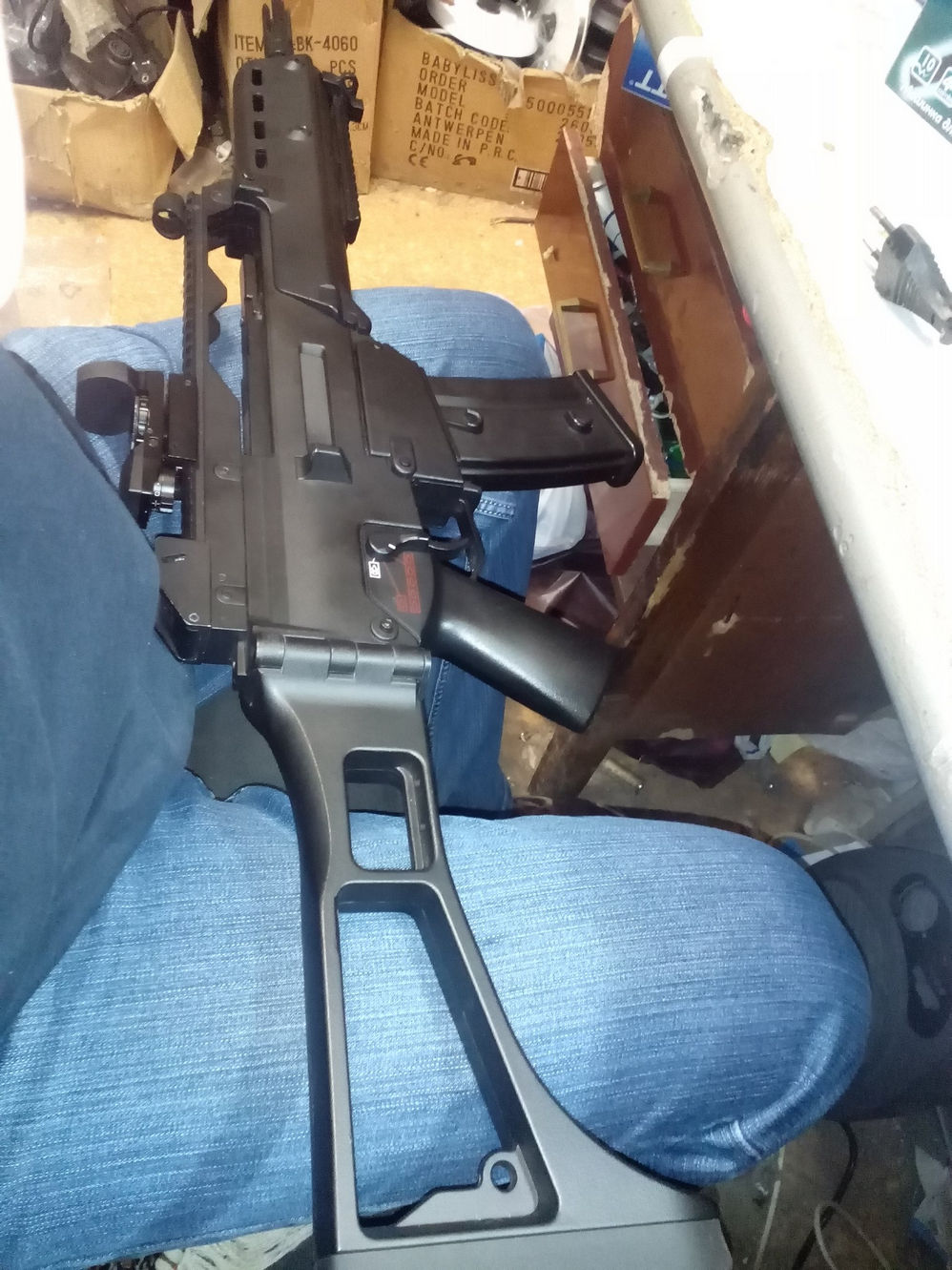 Чёрный страйкбольный привод типа G36 лежит на коленях, вид сбоку — рукоятка, магазин, ствол со складным прикладом