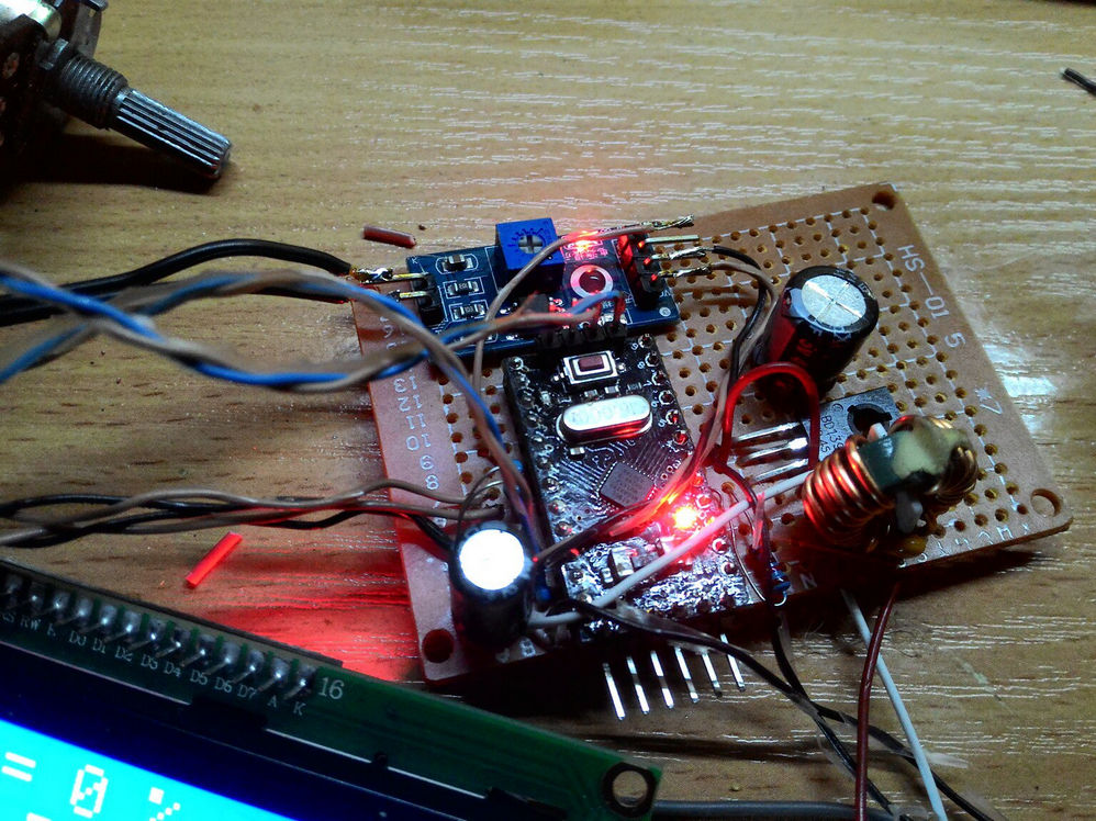 Фото электронной поделки на текстолитовой макетной плате: Arduino Nano (или аналог), плата мотор-драйвера (L298N или аналог) с горящим красным светодиодом-индикатором, конденсаторы и другие компоненты; на переднем плане нижний край синего LCD-дисплея