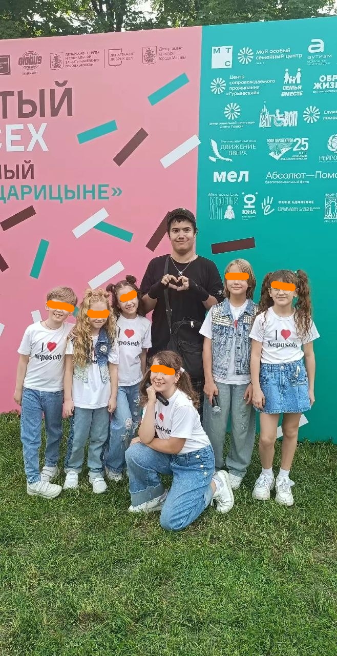 📷 Молодой человек стоит перед баннером «[ОТКРЫ]ТЫЙ [ЧЕ]Х» и «[ОТКРЫ]ТЫЙ [ЦА]РИЦЫНО» в парке Царицыно; рядом — группа девочек в белых футболках «I ❤️»; оранжевые горизонтальные полосы поверх лиц детей.