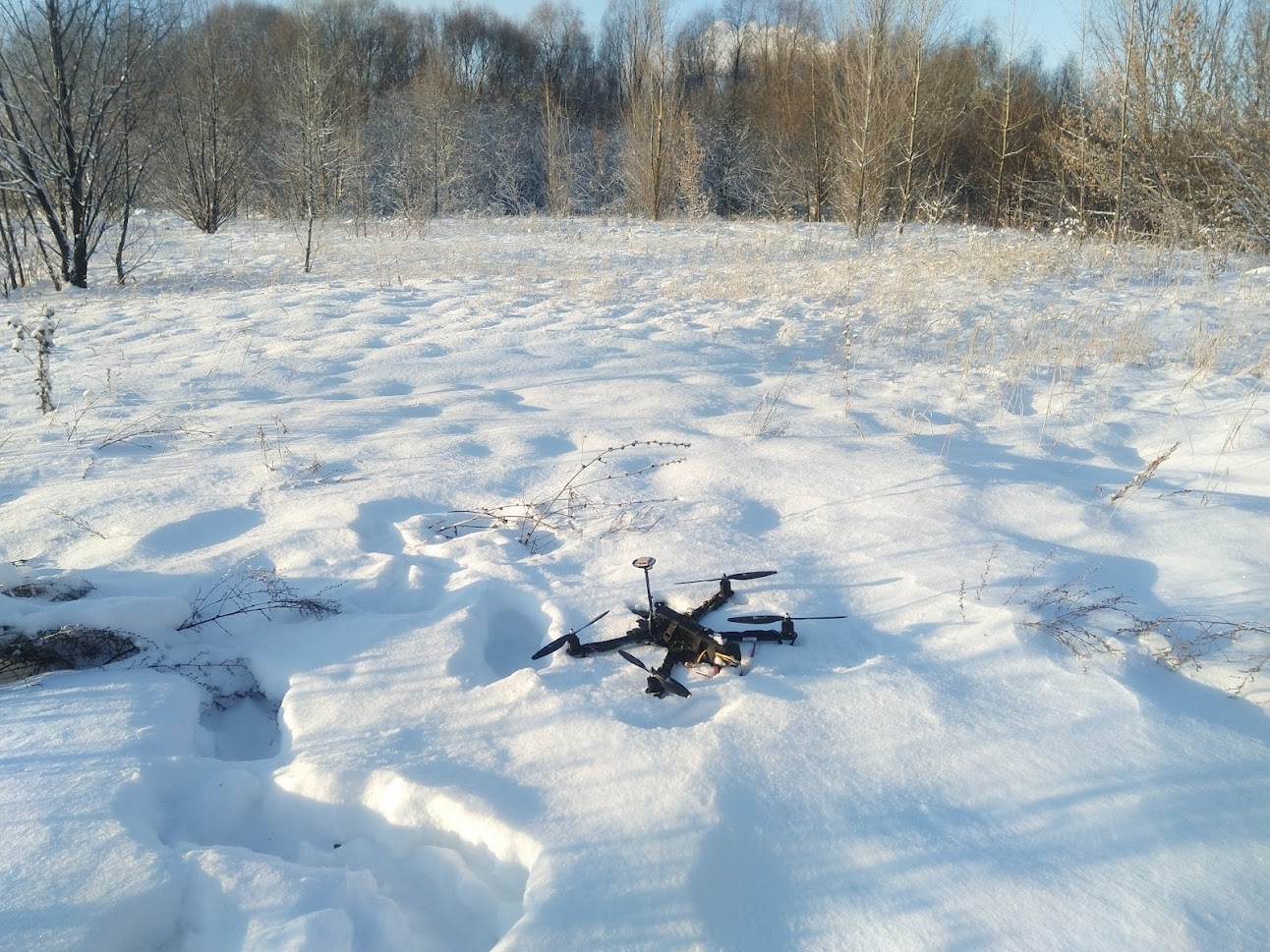 📷 Чёрный квадрокоптер (FPV-дрон) на снегу после посадки: четыре луча с пропеллерами, компактный корпус. Ясный зимний день, глубокий рыхлый снег, тени от дрона. На заднем плане — смешанный лес (берёзы и кустарники без листьев), голубое небо.