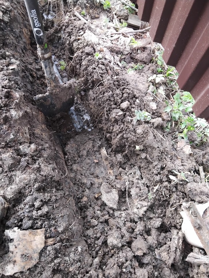 📷 Узкая траншея в тёмном грунте у оранжевого забора, видна лопата — земляные работы.