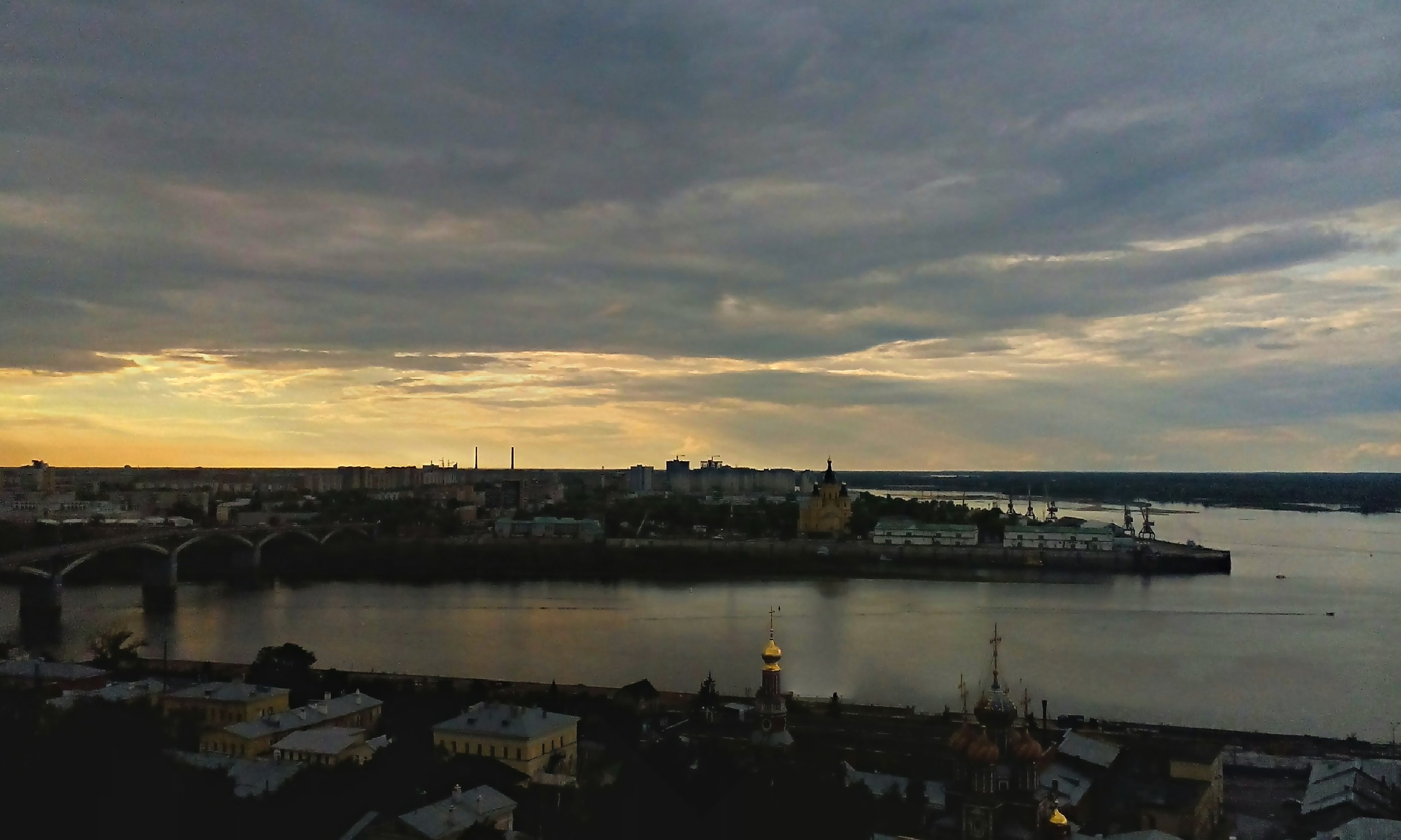 Широкая панорама слияния Оки и Волги в Нижнем Новгороде: собор Александра Невского, портовые краны, закат с золотисто-серыми облаками