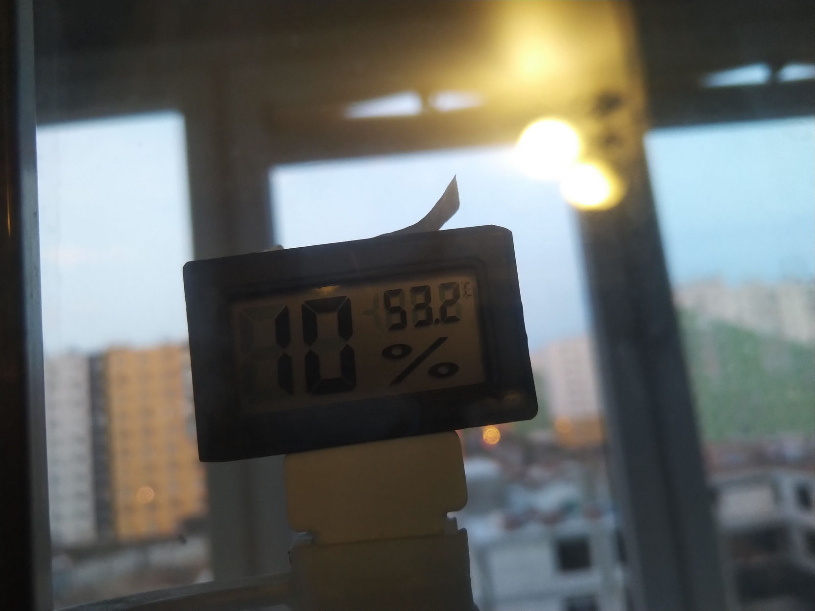 Чёрный термогигрометр прикреплён к стеклу лоджии: дисплей показывает температуру 10°C и влажность 53.2%; за стеклом — вечерний городской пейзаж с многоэтажными домами и зеленью деревьев, справа яркий источник света