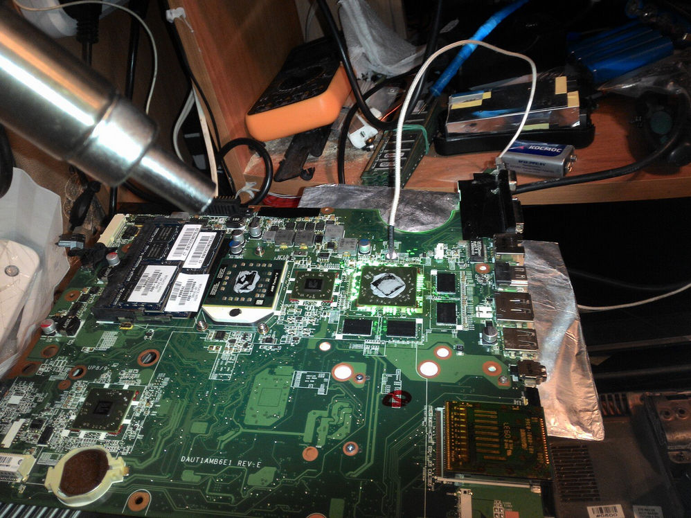 Материнская плата ноутбука DAUTI4MB6E1 REV:E на рабочем столе: слева наведено сопло термовоздушной паяльной станции для рефлоу GPU; сзади оранжевый мультиметр, батарейки «КОСМОС», провода