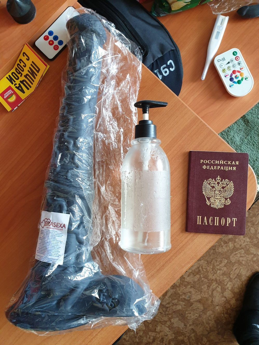 📷 На столе: большой чёрный фаллоимитатор в прозрачной упаковке, флакон лубриканта с помпой, российский паспорт (обложка с гербом); стикеры на стене позади.