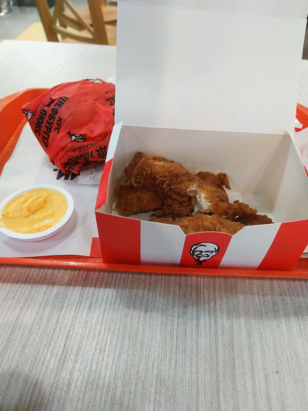Красно-белая коробка KFC с жареными куриными кусочками и ножками; рядом соус в стаканчике и свёрток