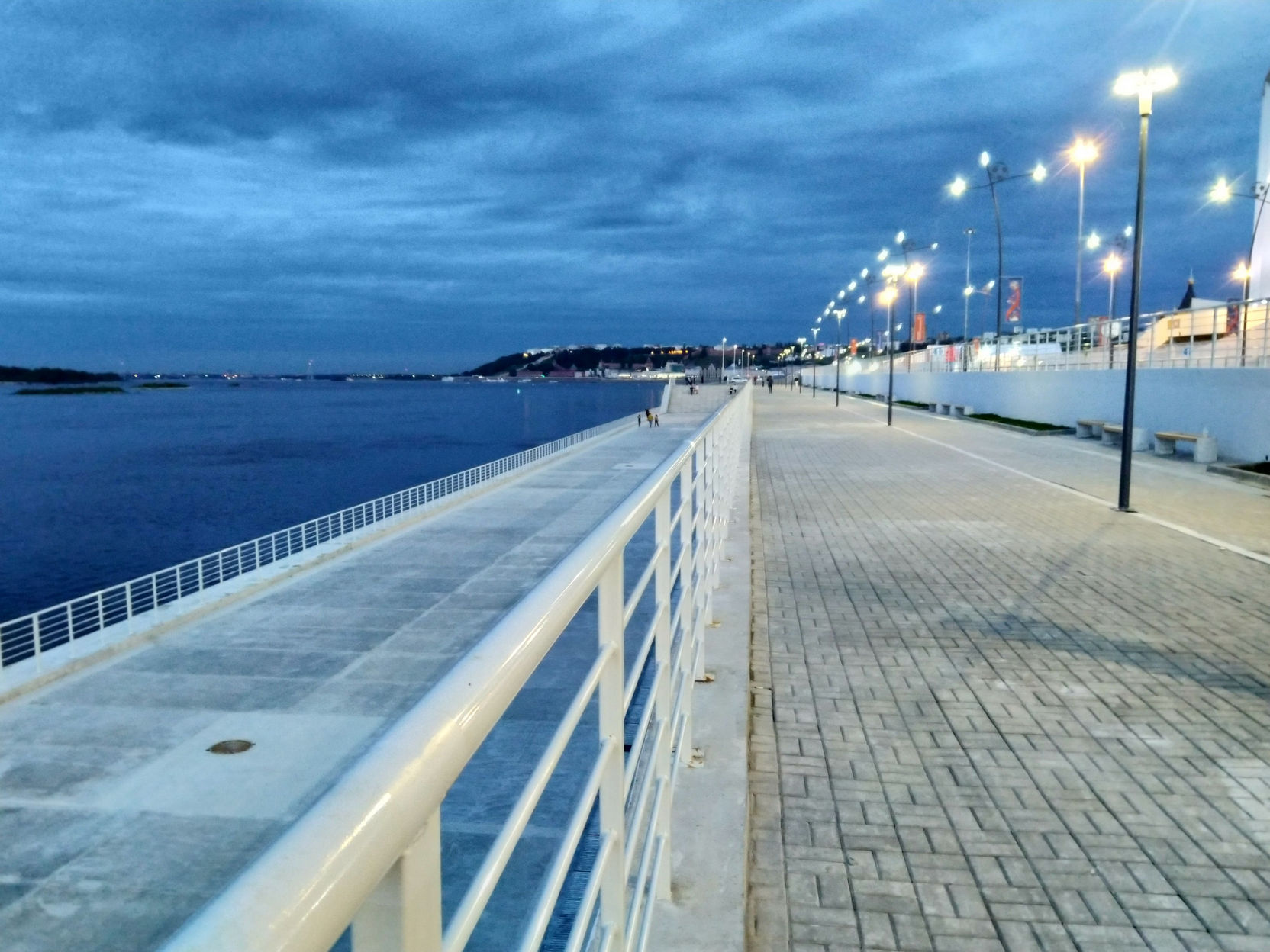 Ночная набережная Нижнего Новгорода: широкий тротуар из светлой плитки с белым металлическим ограждением вдоль Волги; цепочка фонарей уходит вдаль; справа — белое строение; тёмно-синее облачное небо