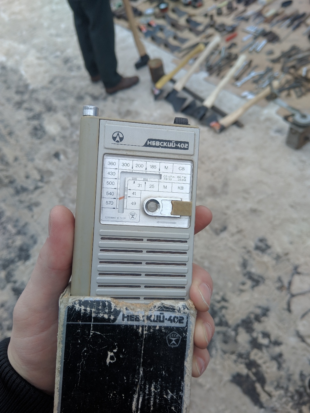 Рука держит советскую портативную радиостанцию «НЕВСКИЙ-402» — серый вытянутый пластиковый корпус с рядом кнопок и антенной, надпись «НЕВСКИЙ-402» на лицевой панели; на заднем плане — барахолка/блошиный рынок с разложенными на земле инструментами и силуэтами людей