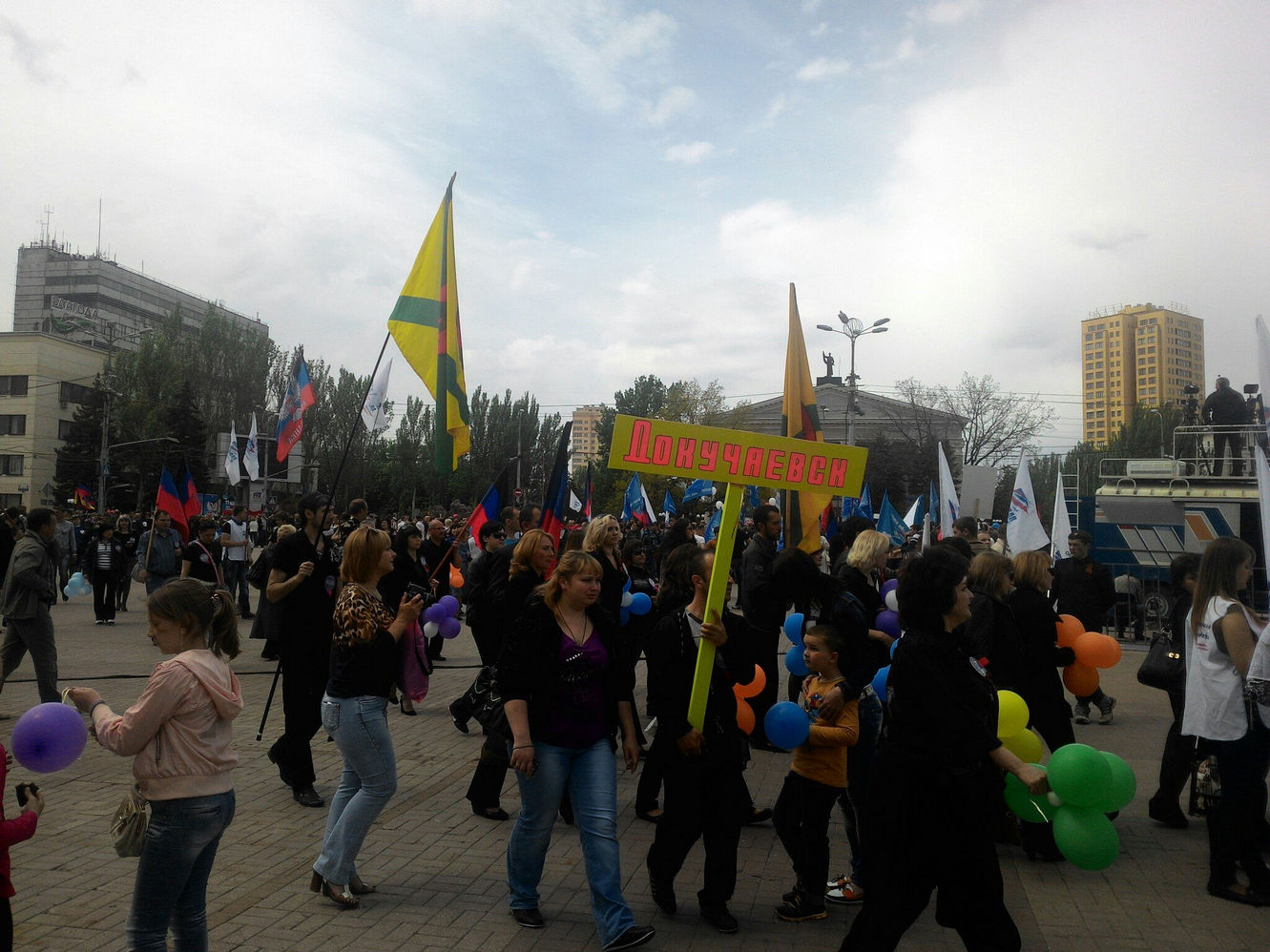 Колонна участников митинга из Докучаевска: женщина несёт табличку «Докучаевск», рядом флаг ДНР и жёлто-зелёный флаг; прохожие с разноцветными шарами; на заднем плане площадь с монументом