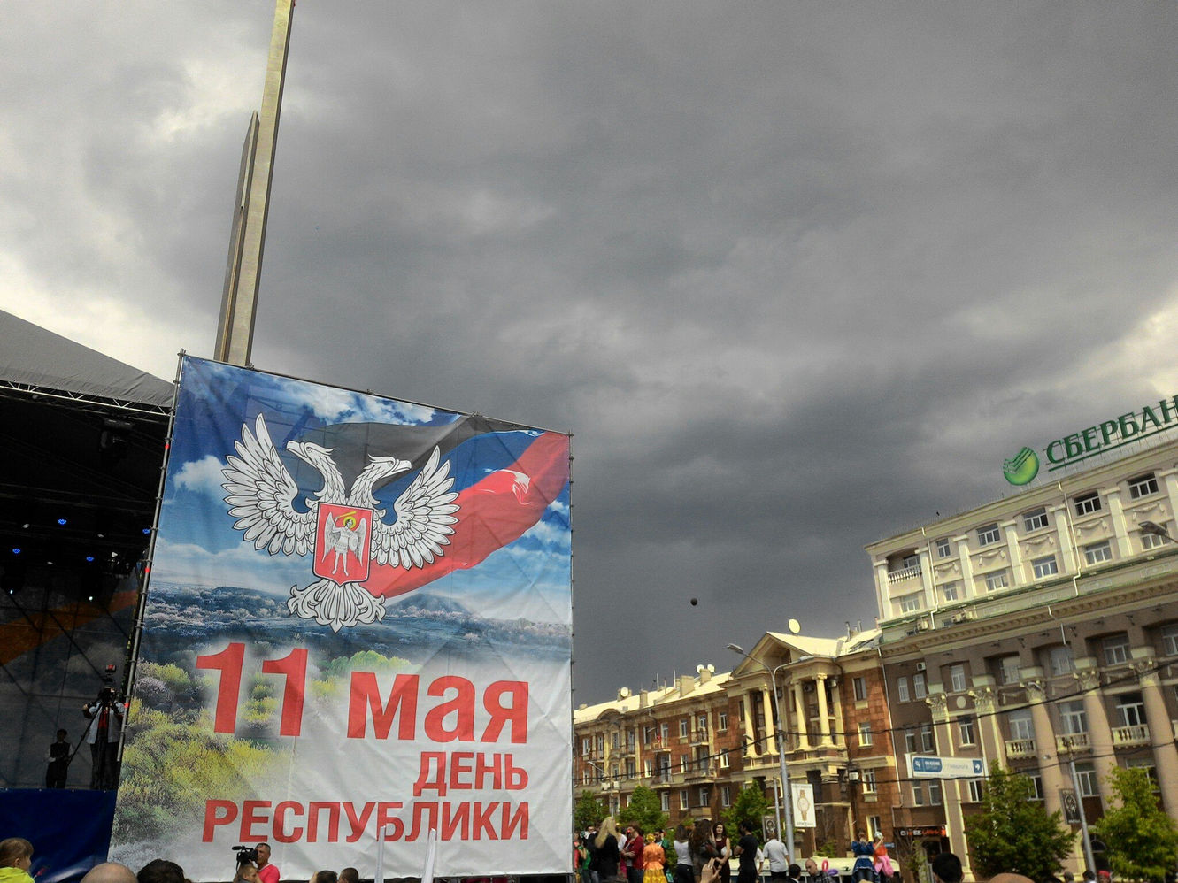 Площадь Ленина в Донецке, 11 мая 2015: праздничный баннер «11 мая — День Республики» с гербом и флагом ДНР у сцены; рядом флагшток и здание со Сбербанком России; нависают грозовые тучи