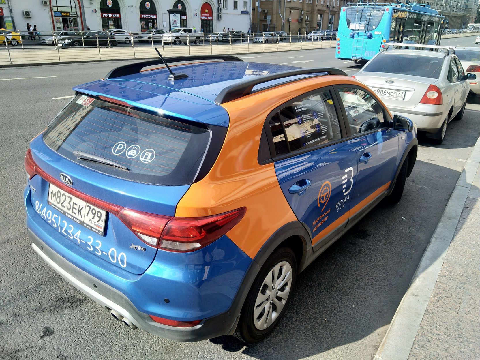 Автомобиль KIA Rio X-Line в сине-оранжевой раскраске Яндекс.Такси, снятый сзади-сбоку: номер МК23ЕК799, логотип сервиса на кузове, номер телефона +7(495)234-33-00; на заднем плане — городская улица с троллейбусом