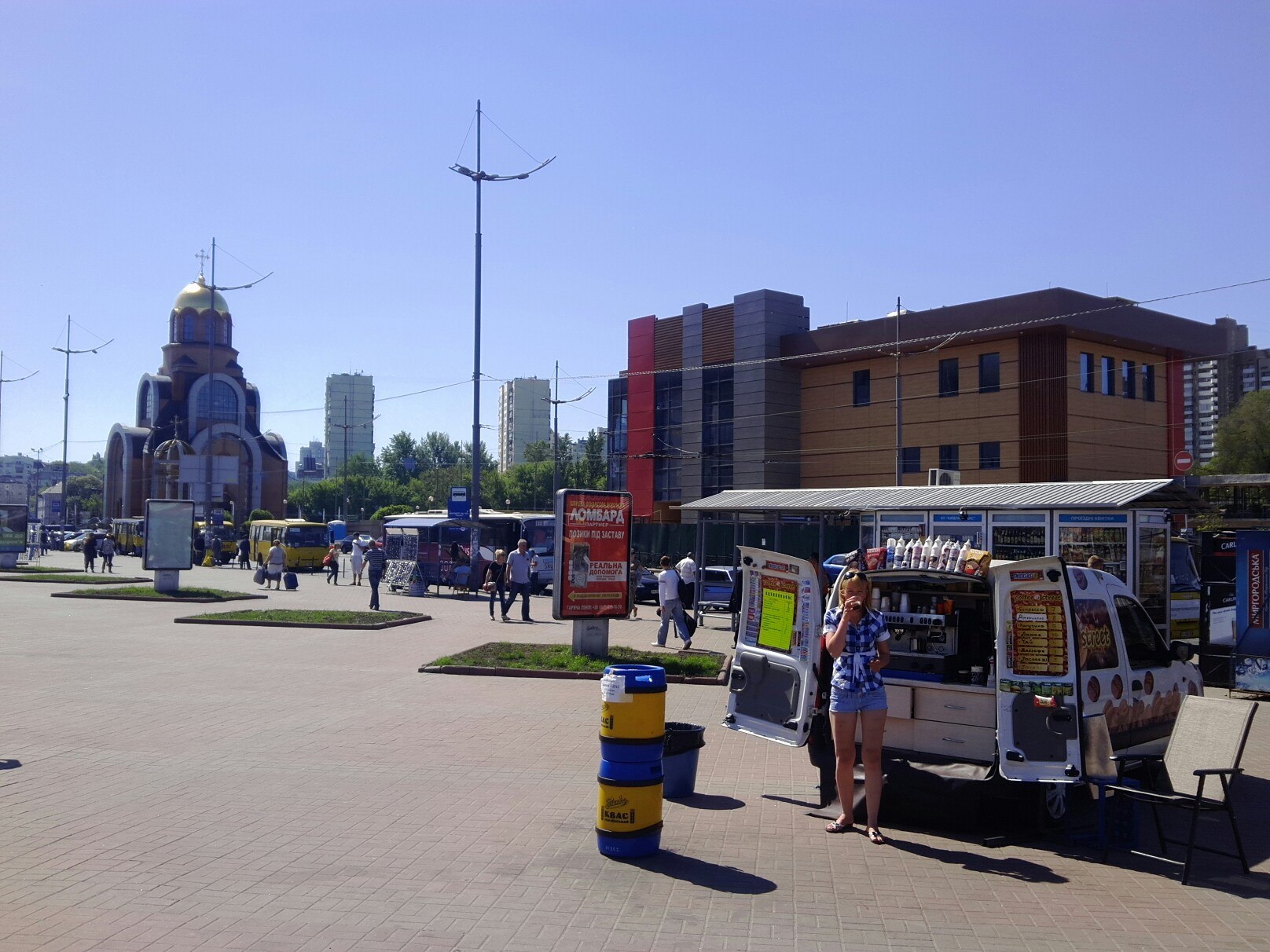 Городская площадь в солнечный день: слева церковь с золотым куполом, в центре мобильный кофейный киоск, прохожие на брусчатке