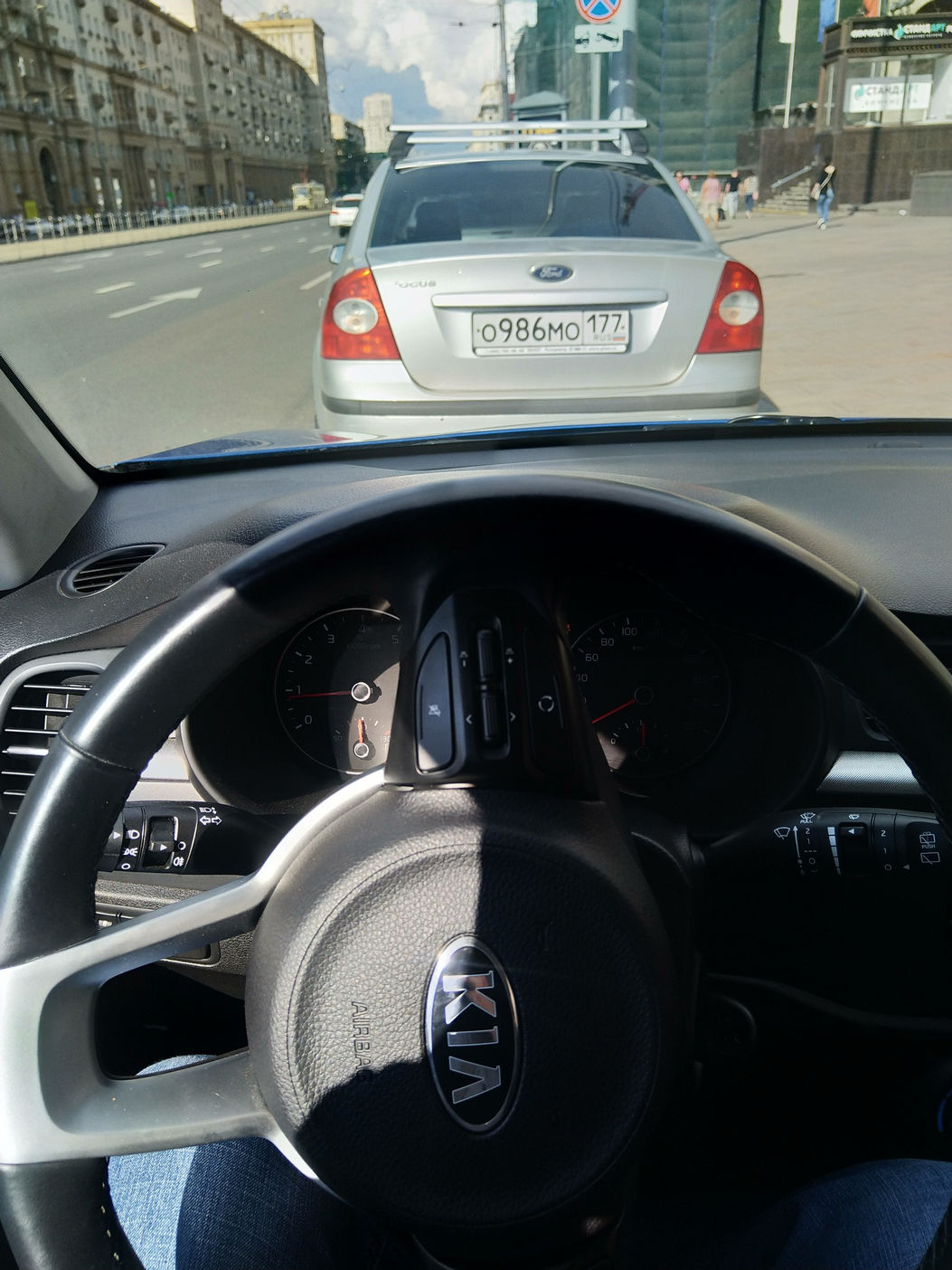 Вид из салона автомобиля (руль с логотипом KIA): впереди на городской улице белый Ford Focus с номером О986МО177, позади него — ещё один автомобиль; многоэтажные здания, ясное солнечное небо