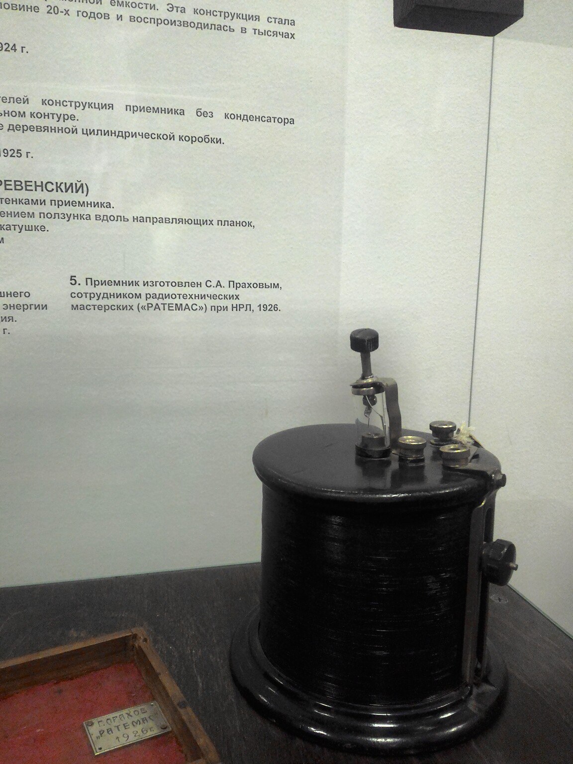 Музейный стенд: чёрный цилиндрический радиоприёмник 1920-х годов (высокий металлический корпус с ручками настройки) на столе рядом с деревянной шкатулкой; на стене — описание: «Приёмник изготовлен С. А. Прозоров, сотрудником радиотехнических мастерских... НРЛ 1926»
