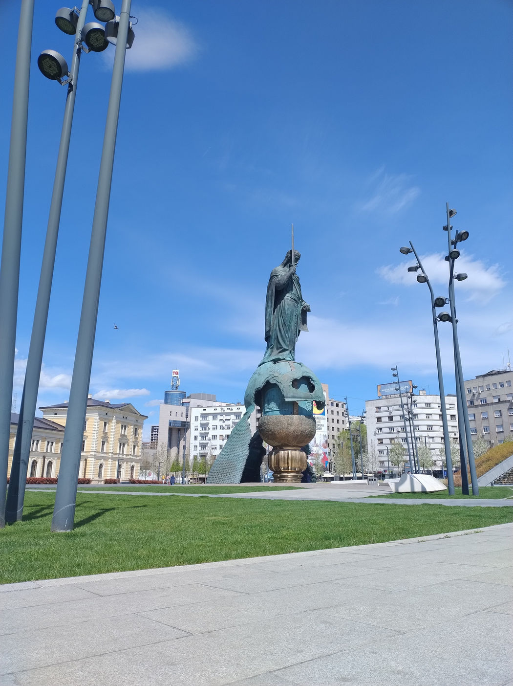 📷 Бронзовая статуя женщины в мантии на шаровом постаменте на городской площади; вокруг жилые и административные здания, голубое небо.