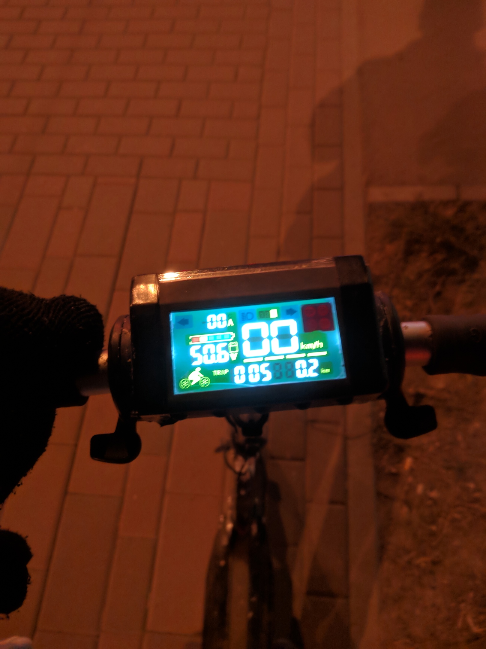 Руль самоката ночью с цветным дисплеем: 0.0A, 50.60V, 00 km/h, TRIP 0.5, 0.2 km; оранжевый уличный свет