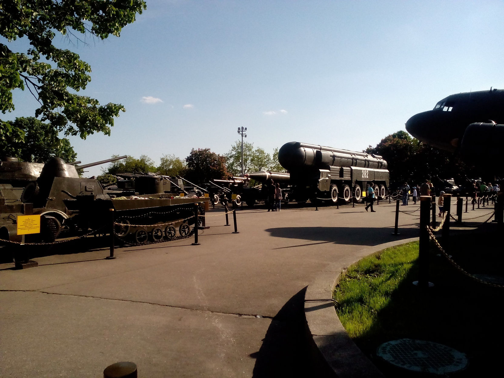 Площадка военной техники: баллистическая ракета на транспортёре, самоходная артиллерия, танки и самолёт; посетители на аллее