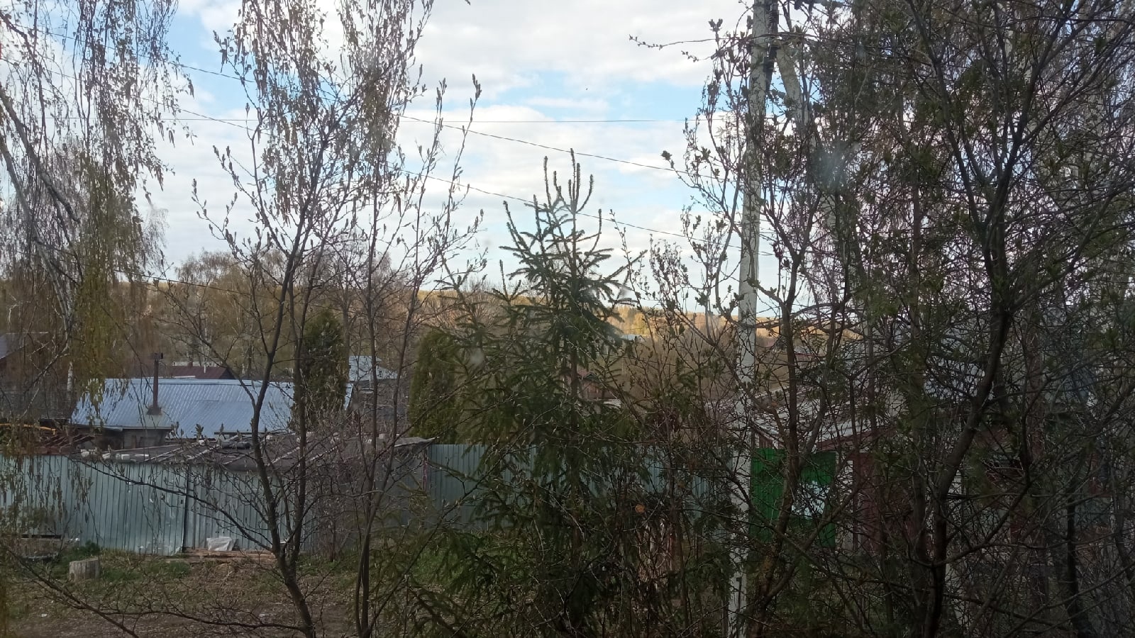 📷 Вид с высоты (окно или балкон) через деревья на водоём в отдалении, ограждения и постройки; частично облачное небо; ранняя весна или поздняя осень.
