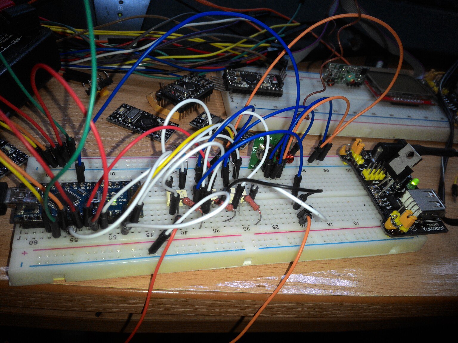 Фото макетной платы (breadboard) на деревянной поверхности: несколько небольших модулей (Arduino Pro Mini или аналоги, оптопары), блок питания MB102 с USB-разъёмом; хаотично проложены разноцветные провода — красные, зелёные, синие, белые, оранжевые, жёлтые