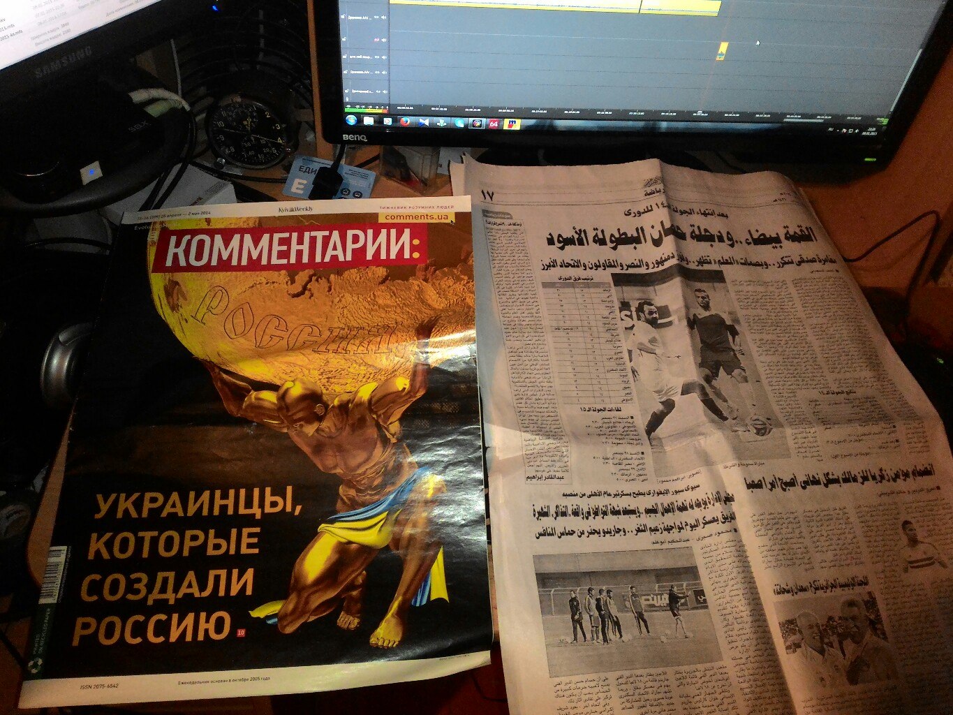 На столе перед монитором лежат два издания: слева — украинский журнал «Комментарии» с обложкой «Украинцы, которые создали Россию» (атлант держит глобус с надписью «Россия»); справа — арабская газета со спортивной полосой о футболе