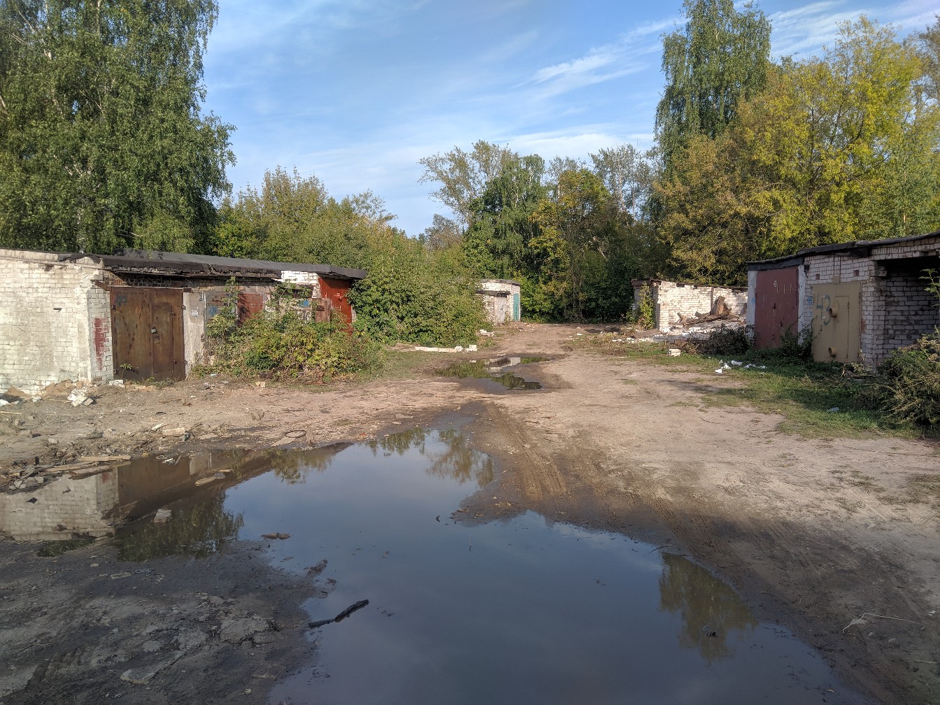 Заброшенный дворик с большой лужей; ряд старых кирпичных гаражей с воротами, деревья, голубое небо