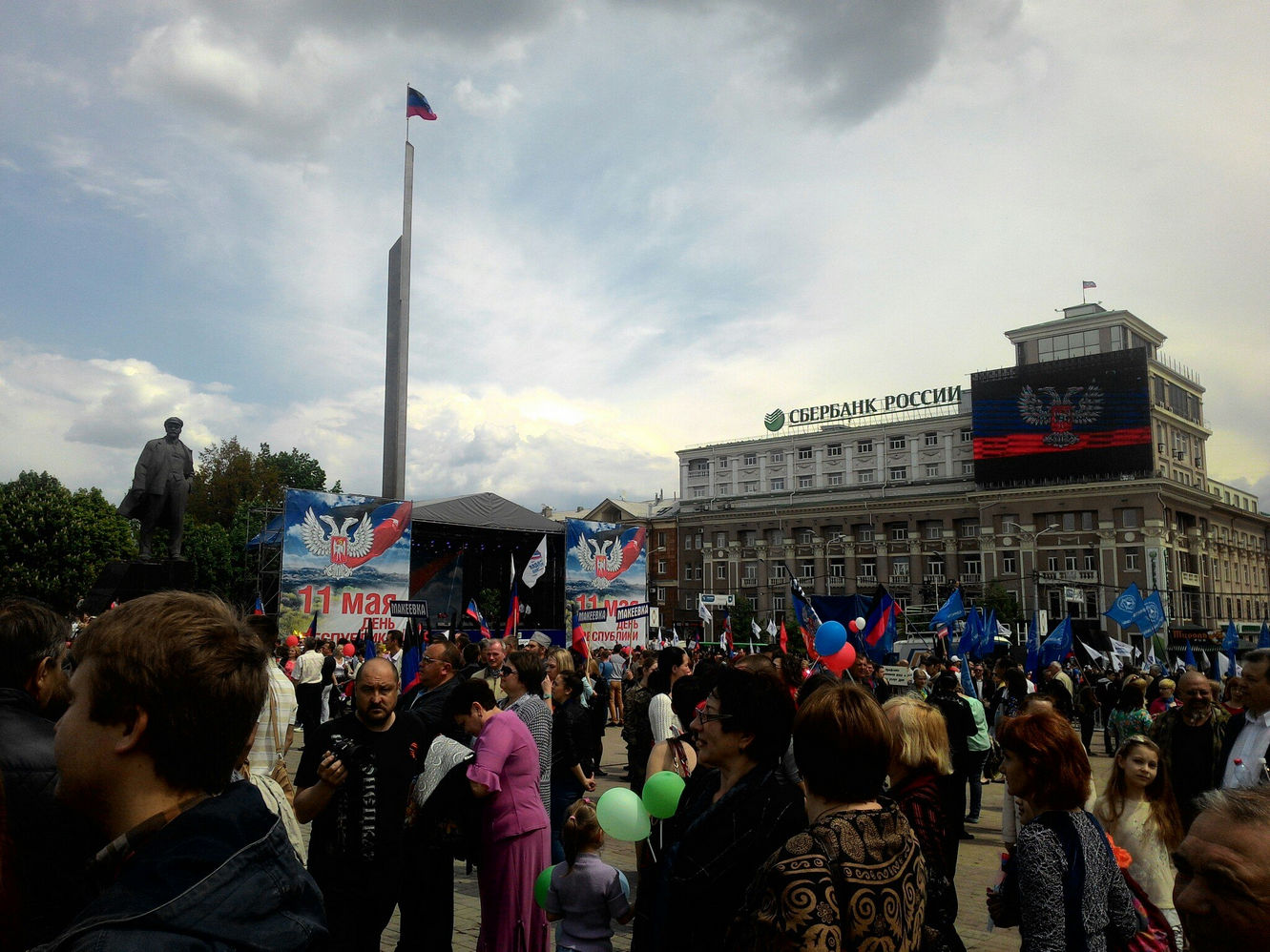Панорама площади Ленина в Донецке во время митинга 11 мая: толпа с флагами ДНР и шарами, сцена с баннерами, флагшток с флагом ДНР; на экране здания Сбербанка России — герб ДНР